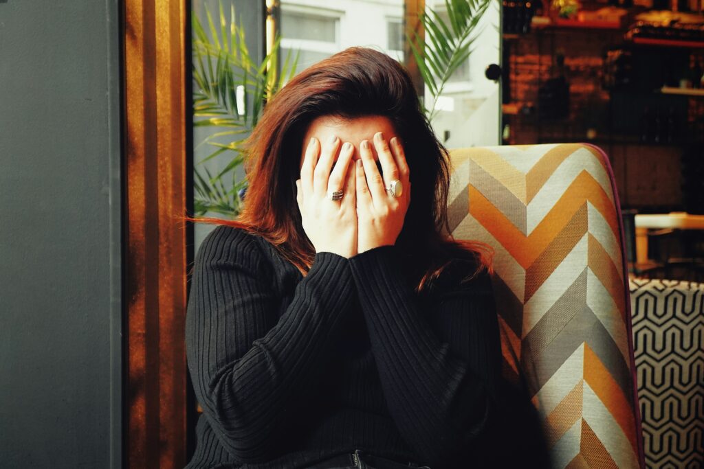 Woman sitting indoors with hands covering their face, wearing a dark ribbed sweater,sitting on a patterned chair.