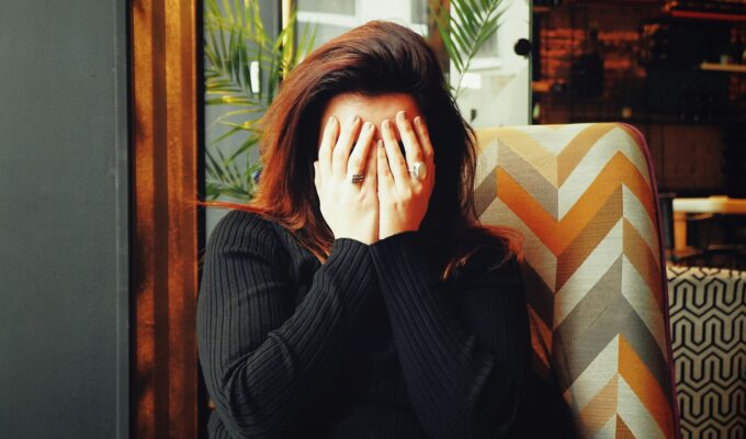 Woman sitting indoors with hands covering their face, wearing a dark ribbed sweater,sitting on a patterned chair.