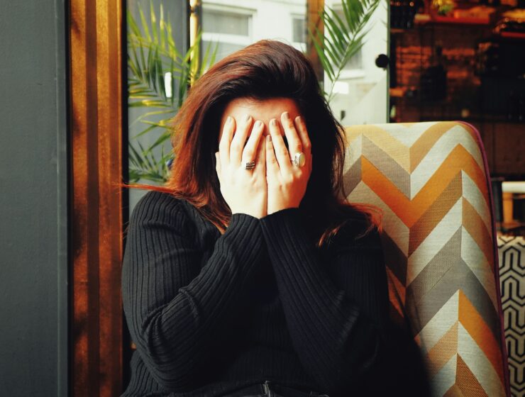 Woman sitting indoors with hands covering their face, wearing a dark ribbed sweater,sitting on a patterned chair.
