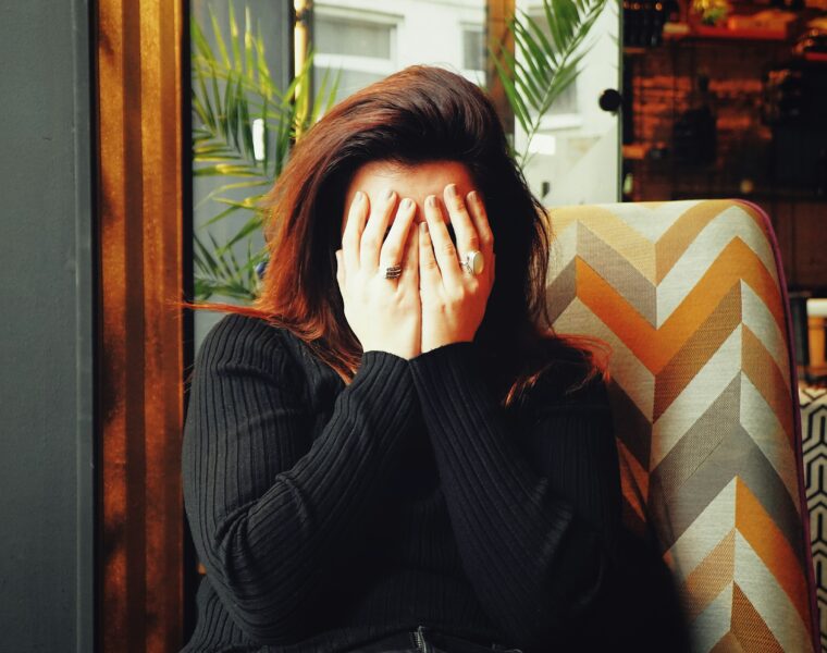 Woman sitting indoors with hands covering their face, wearing a dark ribbed sweater,sitting on a patterned chair.