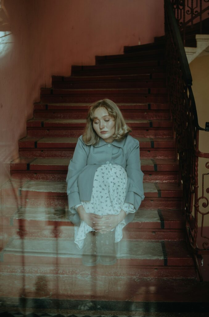 Young woman with blonde hair sits on a red staircase, wearing a light blue coat and a white patterned dress, looking down with hands folded in her lap.
