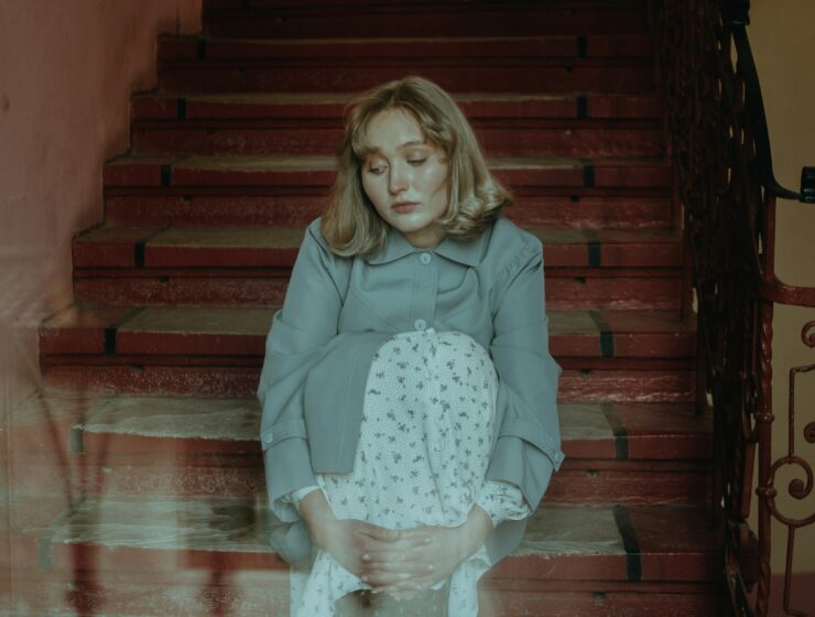 Young woman with blonde hair sits on a red staircase, wearing a light blue coat and a white patterned dress, looking down with hands folded in her lap.