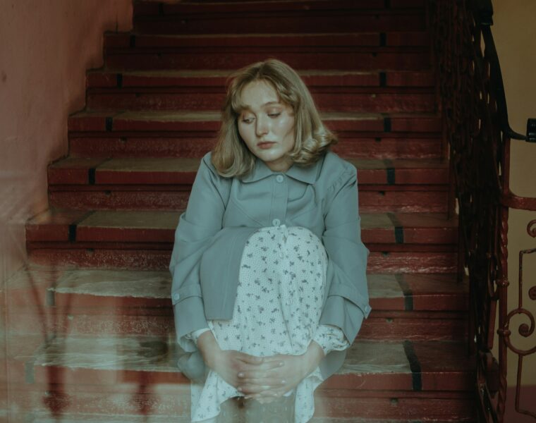 Young woman with blonde hair sits on a red staircase, wearing a light blue coat and a white patterned dress, looking down with hands folded in her lap.